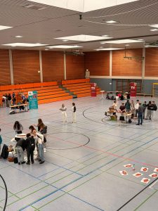 in einer Turnhalle sind mehrere Tische, an denen junge Menschen miteinander reden